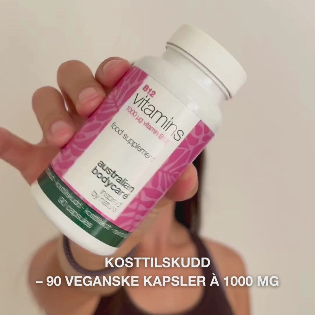 Nærbilde av en hånd som holder flasken med Vitamin B12 kosttilskudd, åpner lokket og heller ut glatte, hvite kapsler—viser produktet for daglig bruk for energi, immunforsvar, nervesystem og redusert tretthet.