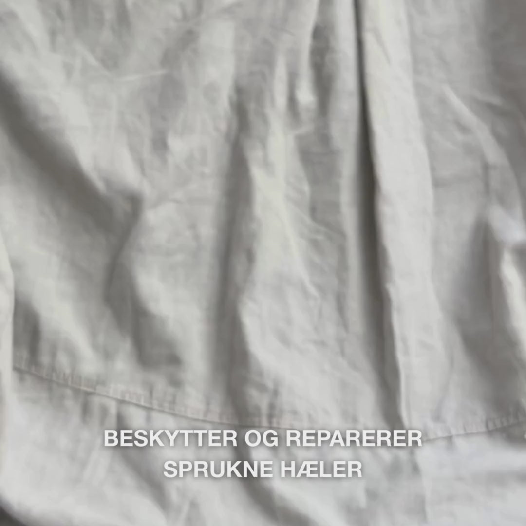 Nærbilde av en hånd som tar en rik, kremet porsjon Heel Repair i håndflaten og masserer den forsiktig inn i tørr, sprukken hud på hælen: den tykke konsistensen absorberes synlig, mykgjør og fukter ru hud, mens 25 % urea, sheasmør og tea tree-olje beroliger og beskytter huden.