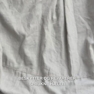 Nærbilde av en hånd som tar en rik, kremet porsjon Heel Repair i håndflaten og masserer den forsiktig inn i tørr, sprukken hud på hælen: den tykke konsistensen absorberes synlig, mykgjør og fukter ru hud, mens 25 % urea, sheasmør og tea tree-olje beroliger og beskytter huden.